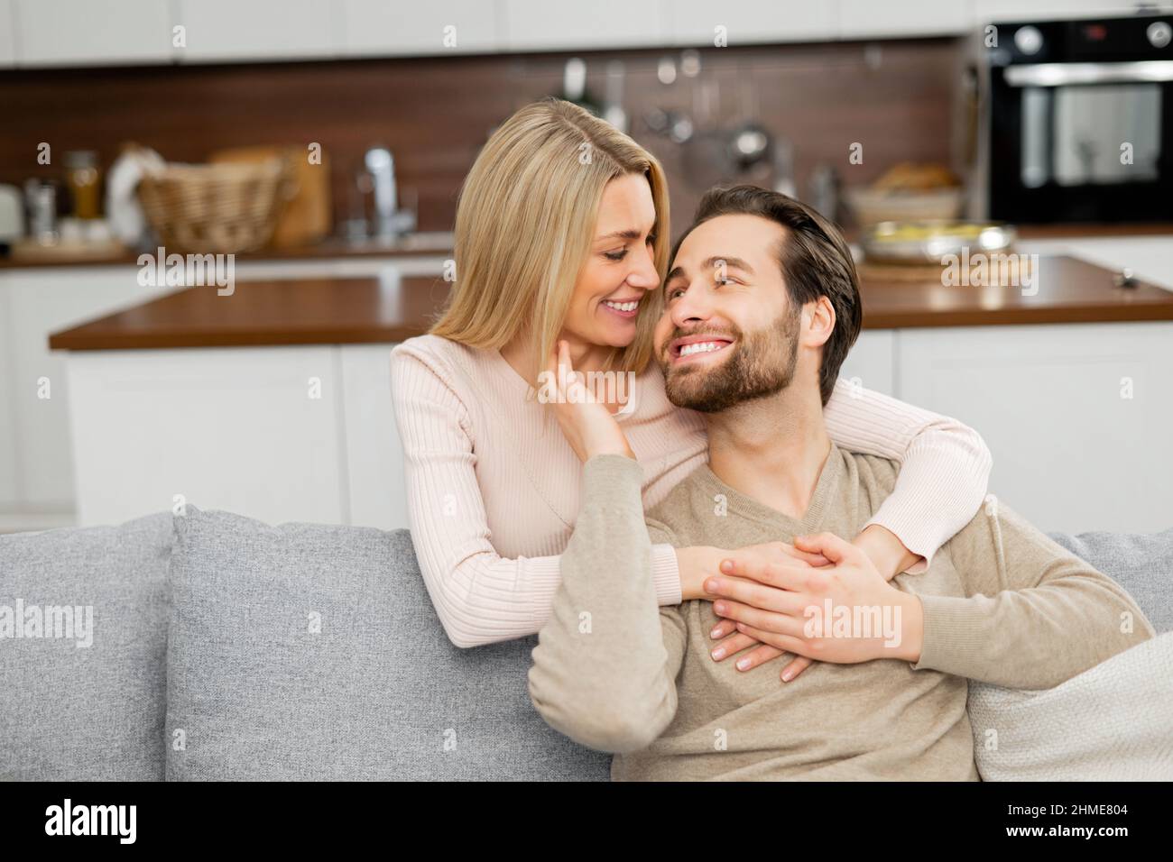 Happy beautiful wife hugging from the back her husband sitting on the couch, they looking at each other and smile. Love and support concept Stock Photo