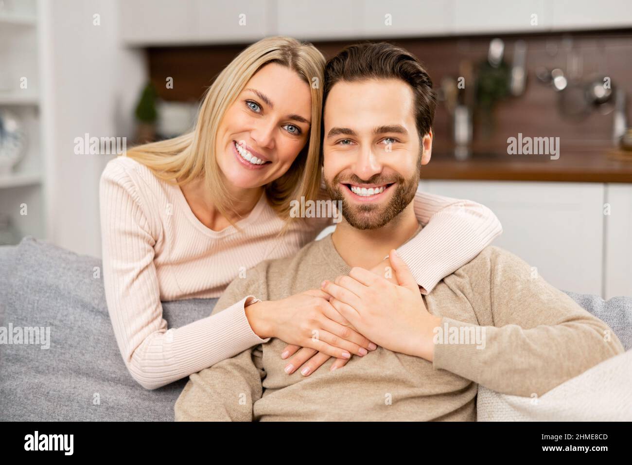 Portrait of lovely couple at home. Handsome man sitting on the comfortable couch, beautiful blonde woman hugging his from the back. Spouses spend time together Stock Photo