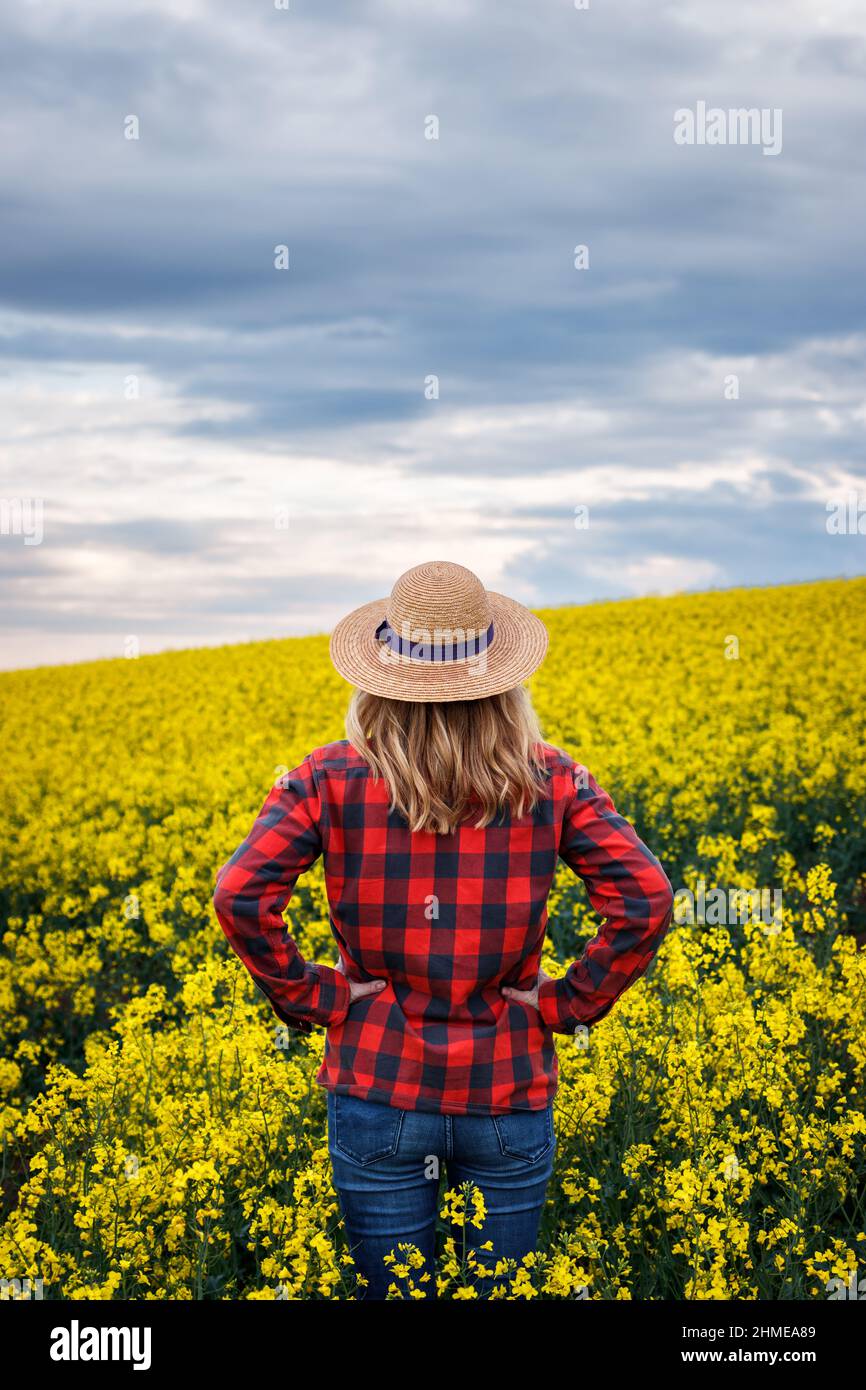 Proud farmer is looking at blooming oilseed field during sunset. Woman with straw hat and plaid shirt standing in rapeseed agricultural field Stock Photo