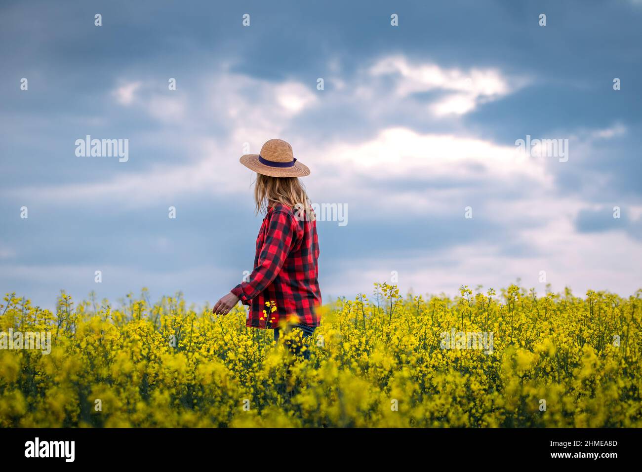 Proud farmer is looking at blooming oilseed field. Woman with straw hat and plaid shirt standing in rapeseed agricultural field Stock Photo