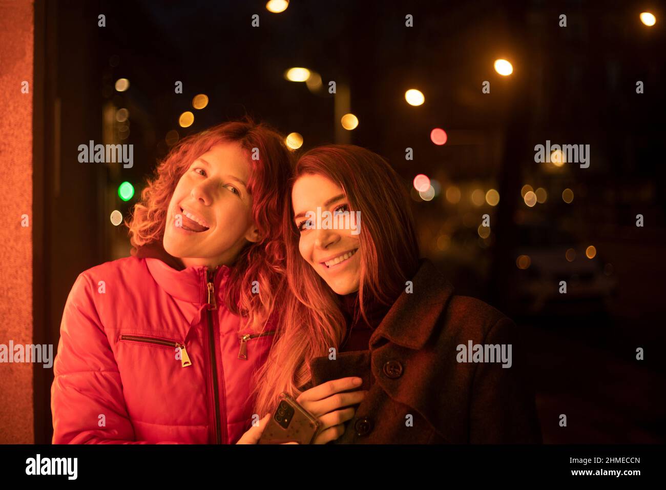 couple of women at night looking at the camera happily illuminated with shop window lights Stock Photo