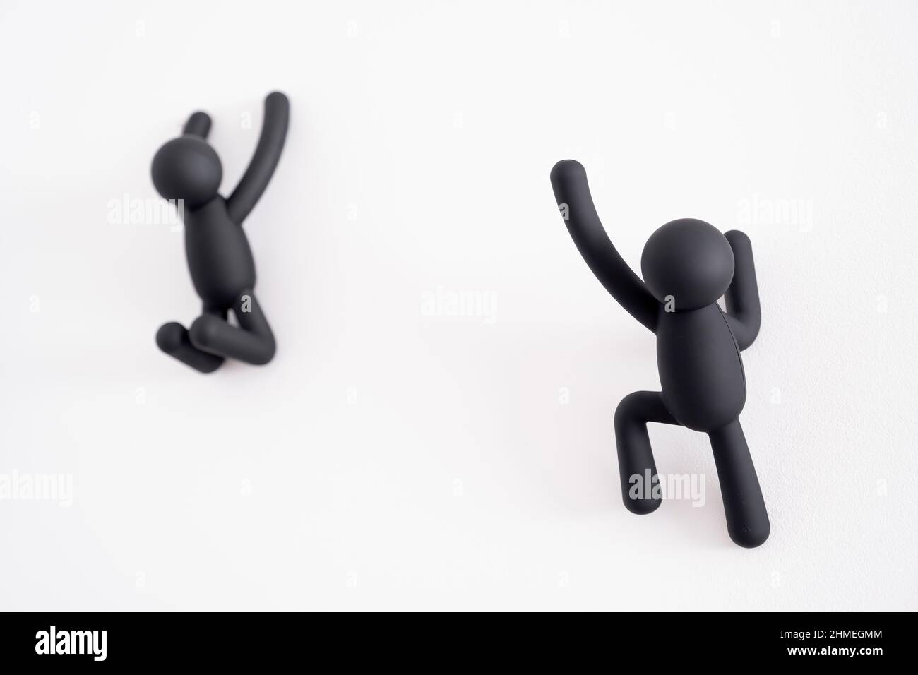 2 black 3D puppets climbing over a white background, human character , person hanging from a white wall, one trying to help the other that is out of f Stock Photo
