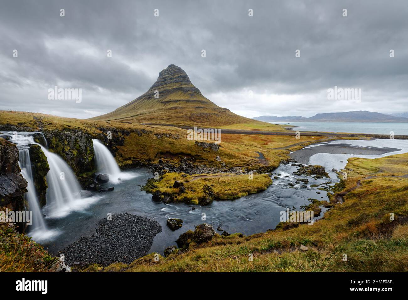 Panoramic view over a wide coastal landscape dominated by the prominent mountain Kirkjufell, in the foreground a waterfall and rapids, above a cloudy Stock Photo