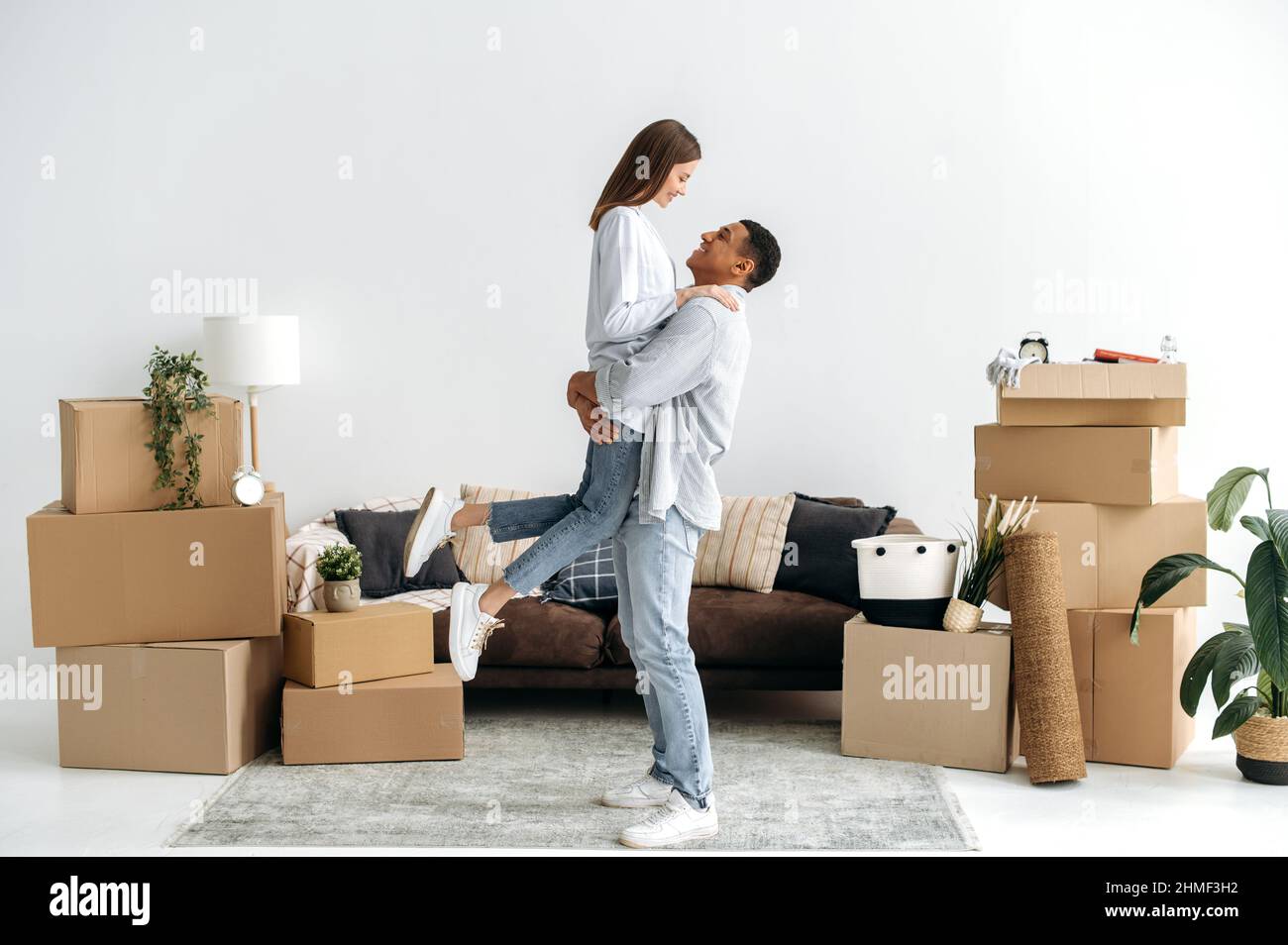 A loving young couple rejoices in their new home. Guy holds his beloved wife in his arms, in living room, they moved to a new apartment, next to cardboard boxes with things. Real estate investment Stock Photo