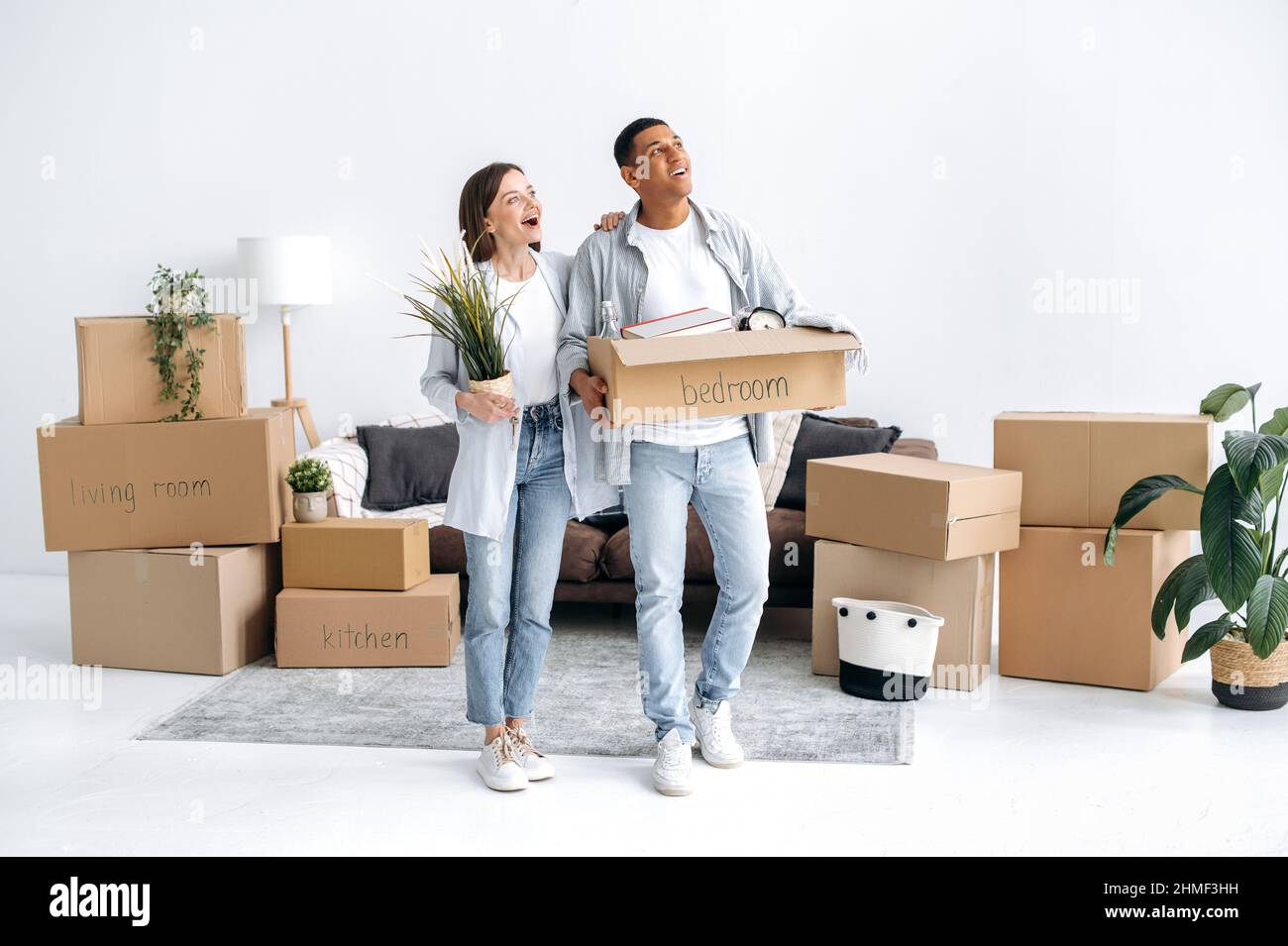 Moving to a new home. Excited couple in love, husband and wife of different nationalities, moved to their new home, guy holds a box with things, girl a flowerpot, enthusiastically inspecting new house Stock Photo