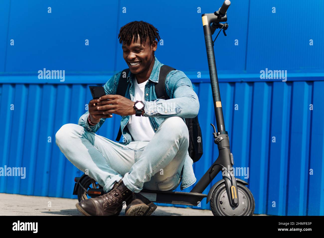 Stylish smiling african american man sitting on his electric scooter near a large building, using a mobile phone while sitting near a blue wall, stude Stock Photo