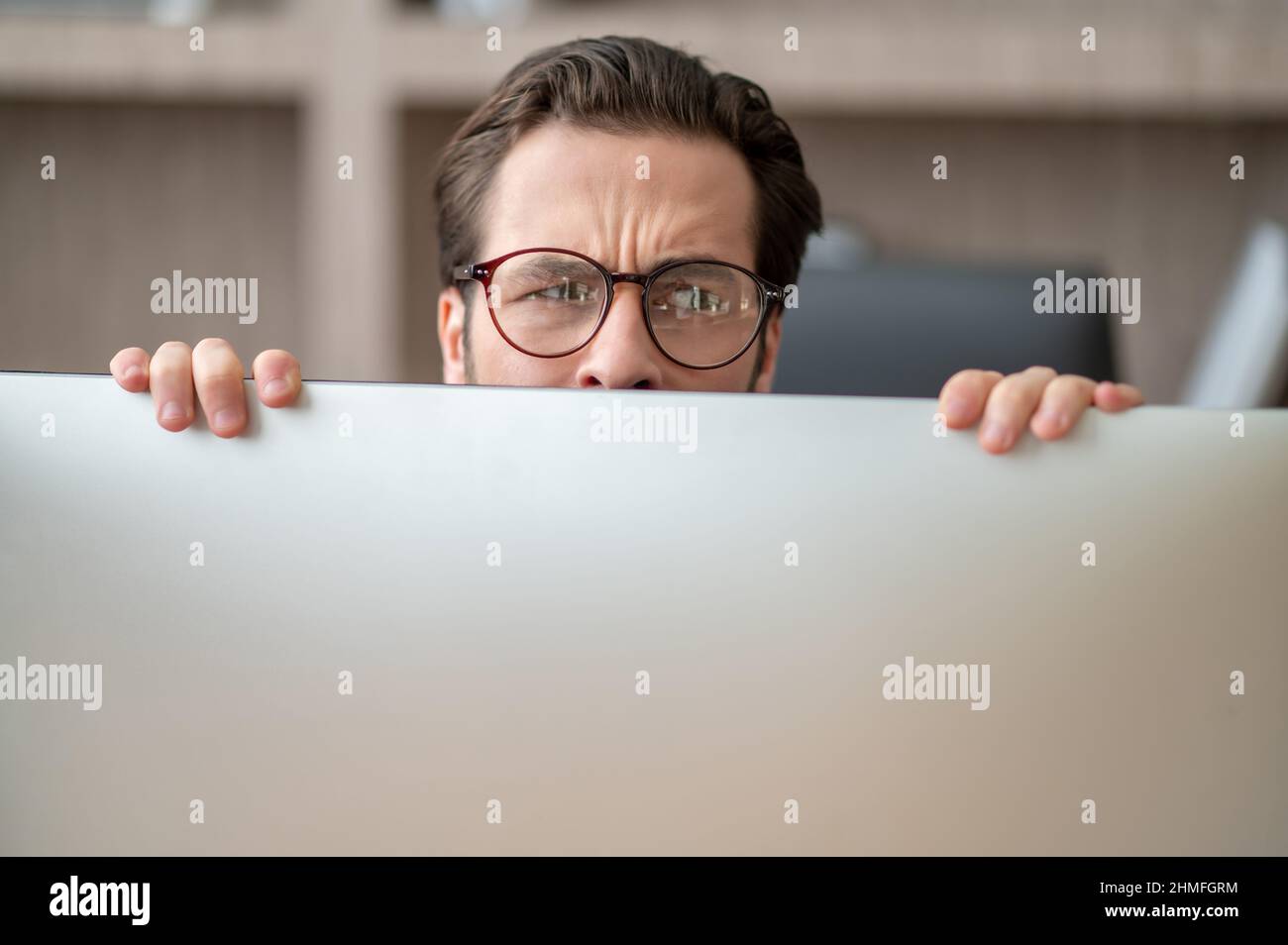 A dark-haired man hiding behind the computer and looking stressed Stock Photo