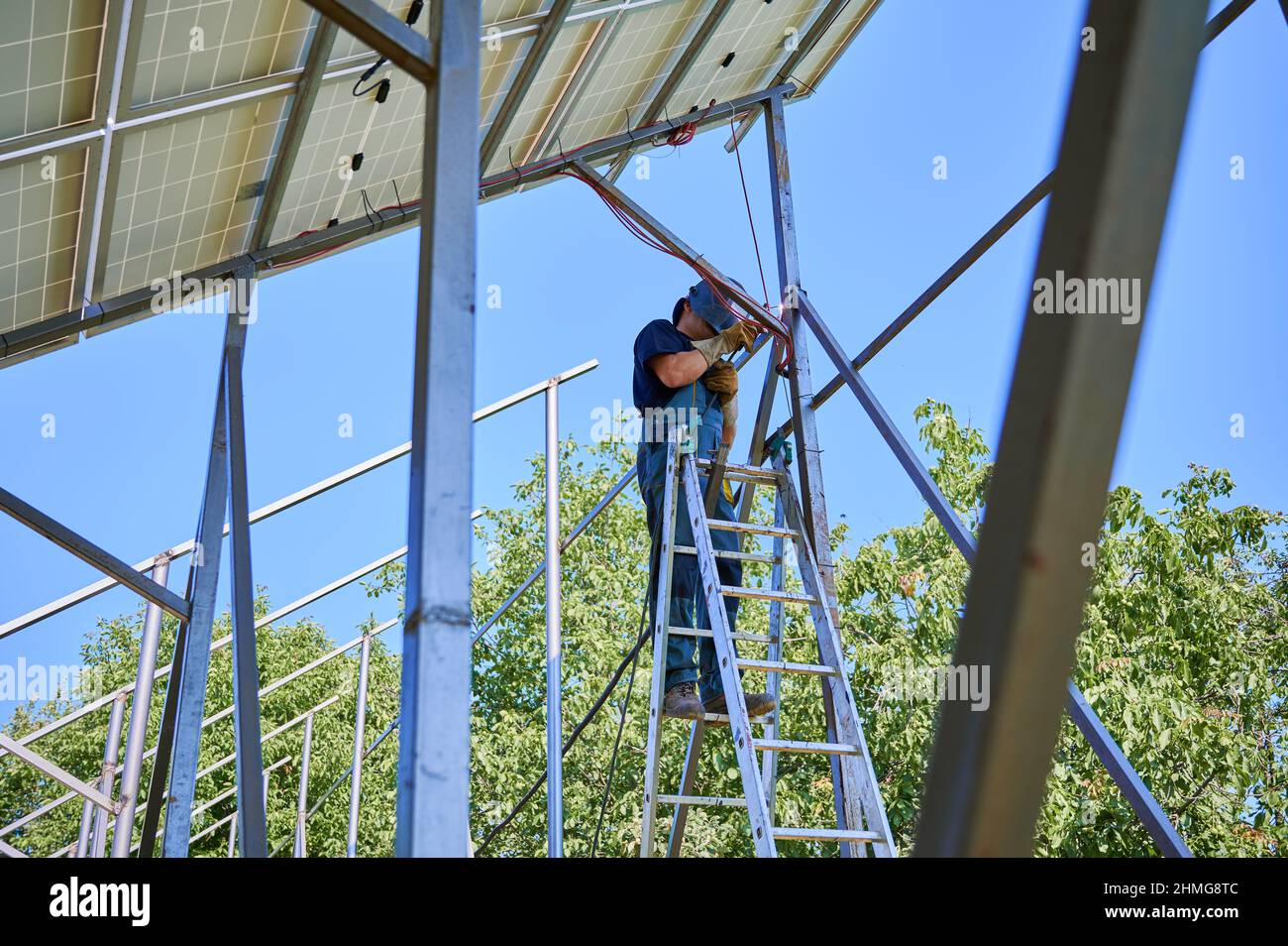 Male worker standing on ladder and welding metal poles of photovoltaic solar system. Man wearing safety welding mask while mounting support structures for solar panels. Stock Photo