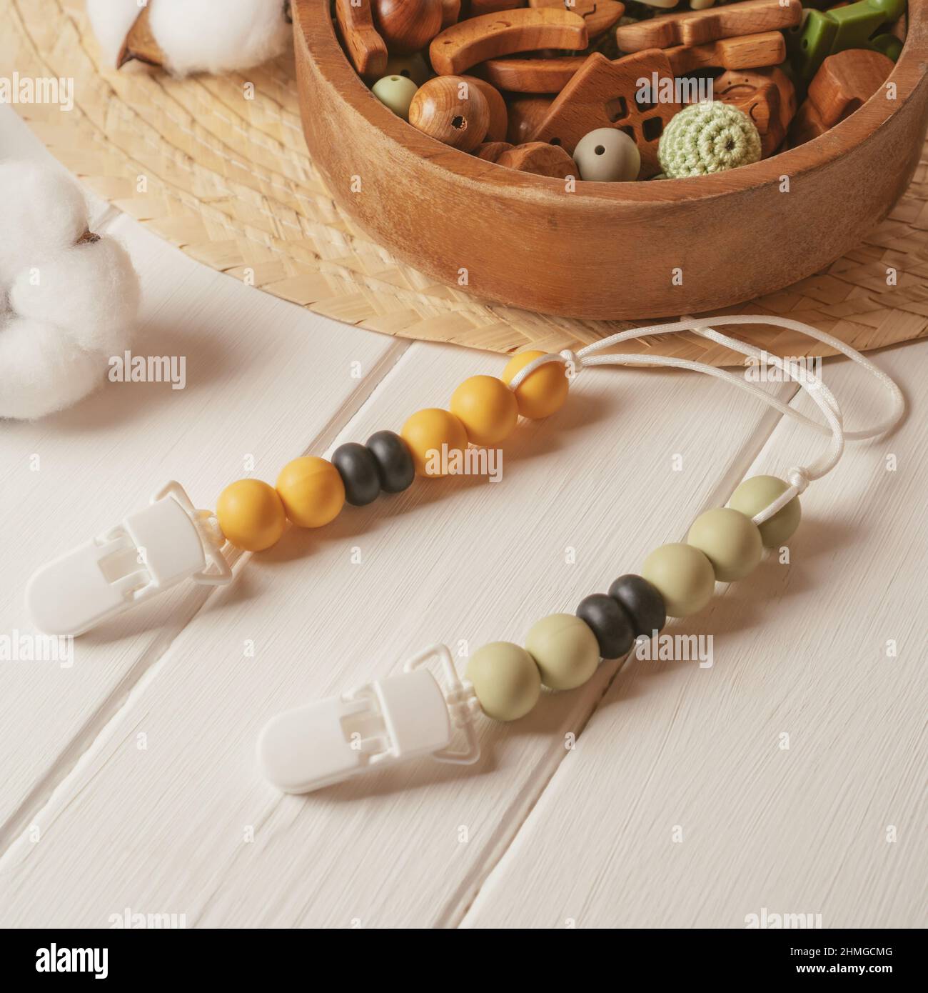 White plastic clothespin with white, black and orange wooden beads on white laces for baby pacifiers near wooden bowl with different beads on white wo Stock Photo
