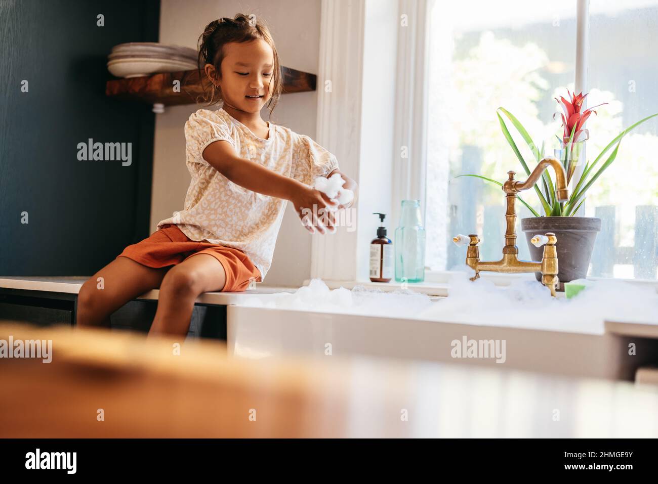 Young girl playing with soap bubbles in the kitchen. Cute little girl having fun with white foam while sitting next to the kitchen sink at home. Stock Photo
