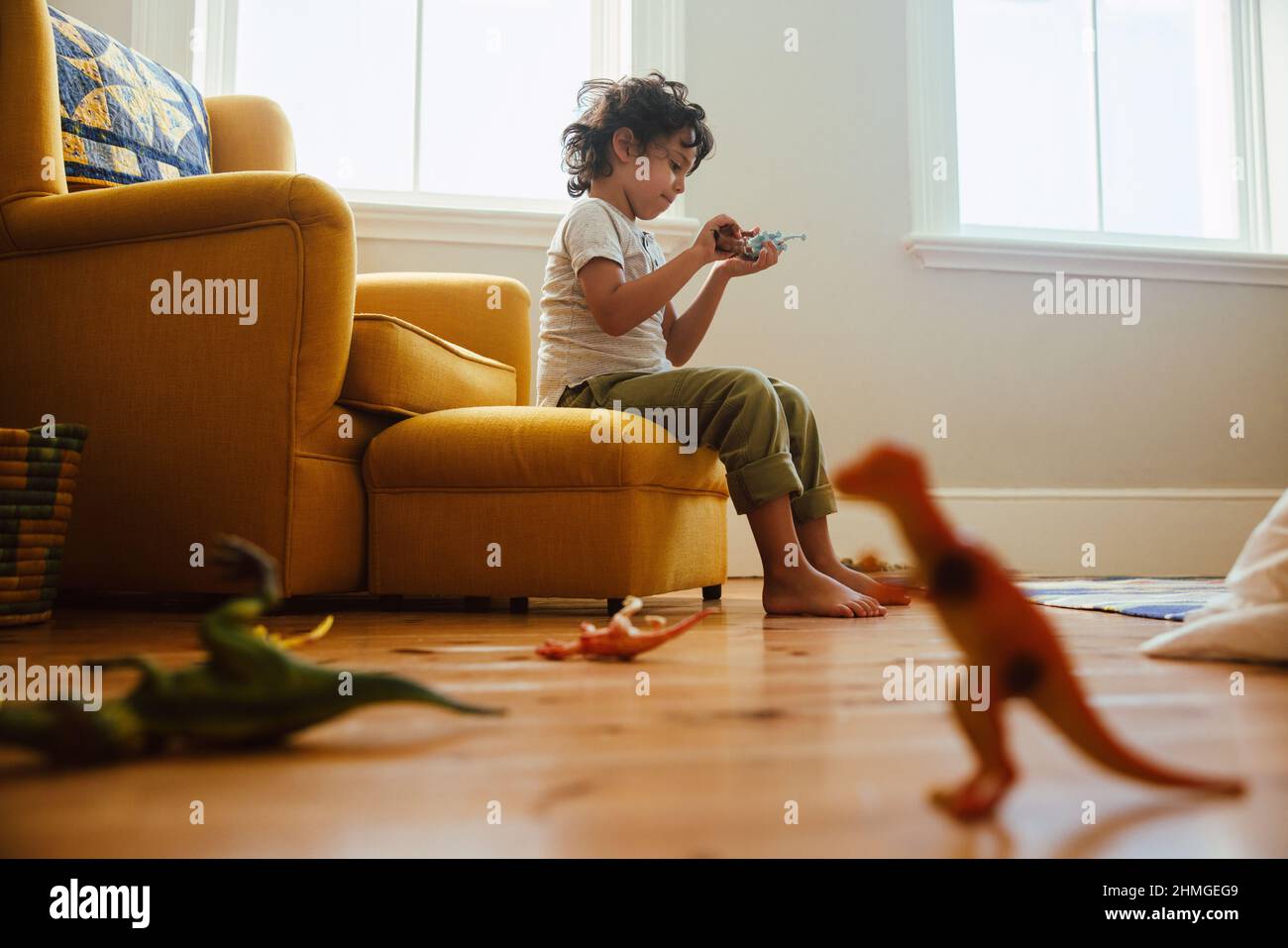 Little boy playing with colourful animal toys in his play area at home. Adorable young boy sitting on a couch while playing a fight game with dinosaur Stock Photo