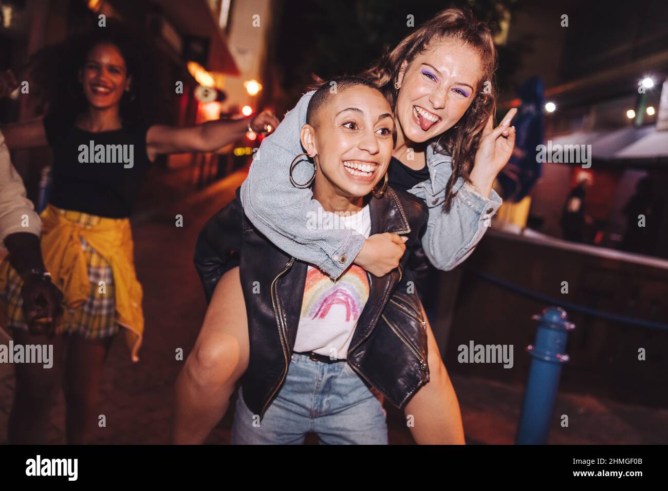 Playful young woman piggybacking her best friend outdoors at night. Happy young women having fun while going out with their friends in the city. Vibra Stock Photo