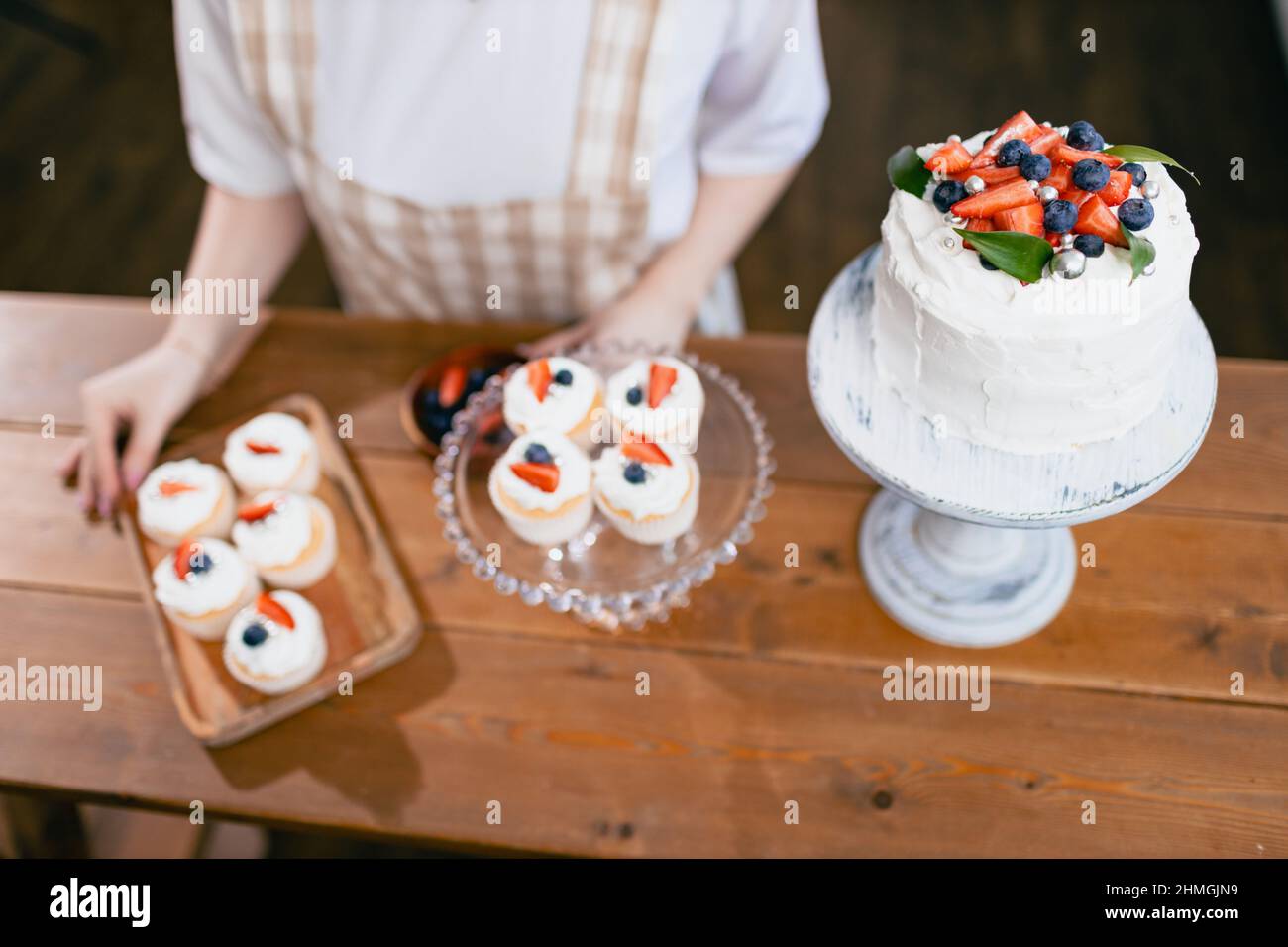 Pastry chef confectioner young caucasian woman decorate cake on kitchen table. Cakes cupcakes and sweet dessert top overhead view Stock Photo
