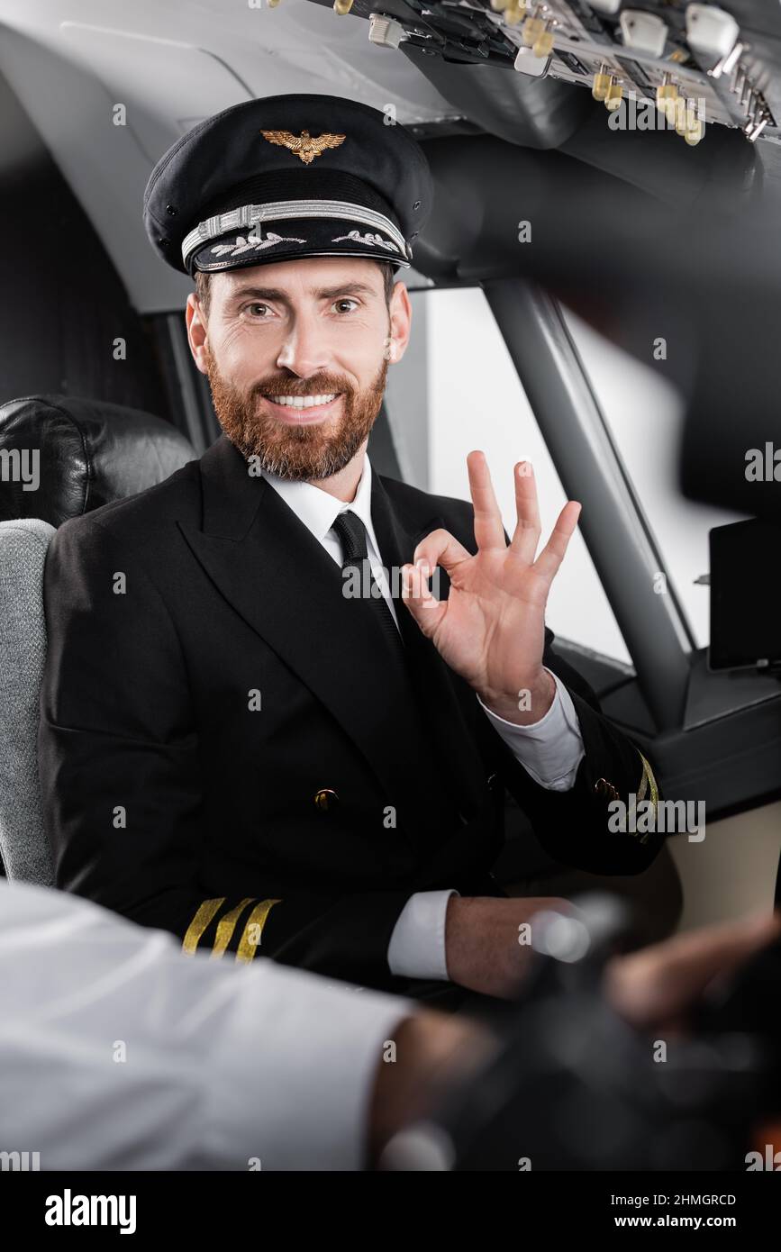 bearded pilot in uniform showing okay and smiling near blurred copilot