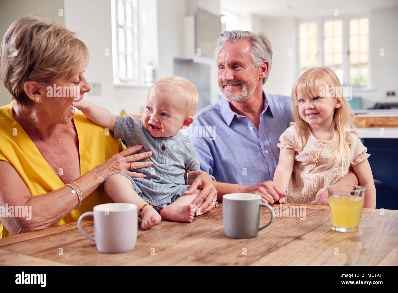 Grandparents Looking After Laughing Granddaughter And Baby Grandson Sitting At Kitchen Table Stock Photo