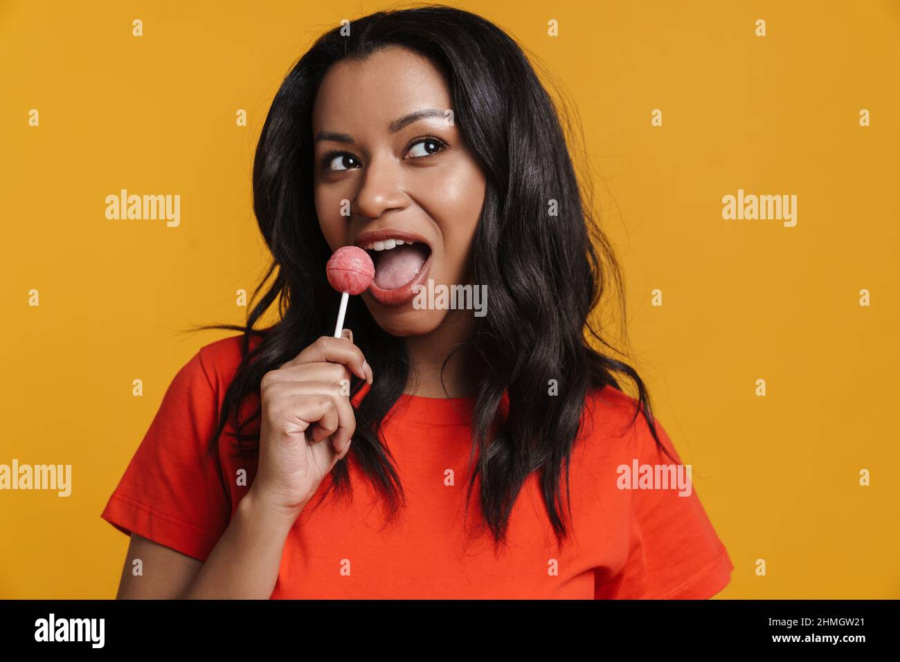 Young black woman smiling while licking lollipop isolated over yellow background Stock Photo