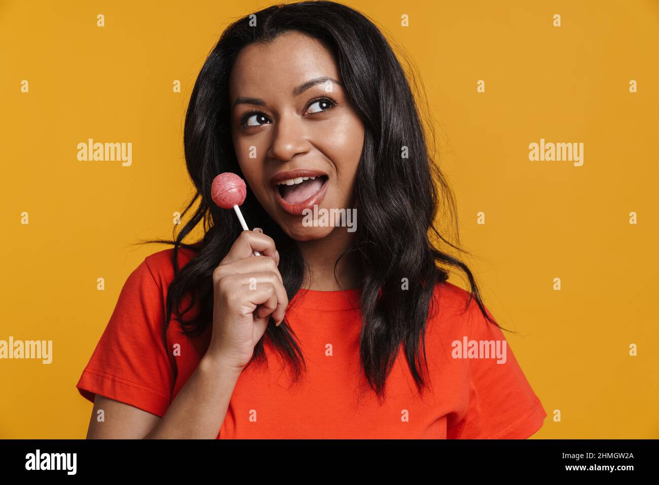 Young black woman smiling while licking lollipop isolated over yellow background Stock Photo