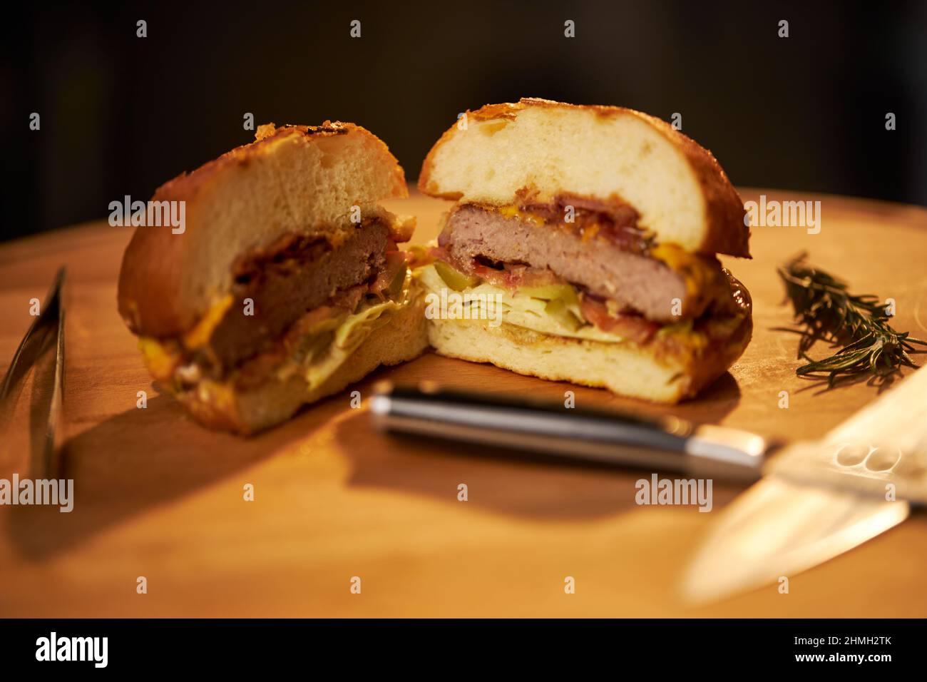 Close up view of grilled beef burger cut in half with knives on cutting board. Juicy homemade cheeseburger with salad, onion and stick of rosemary on wooden board. Concept of fast food.  Stock Photo