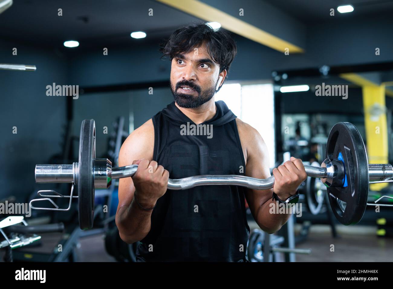 Young Indian bodybuilder busy doing biceps workout by lifting weight using barbells at gym - concept of musclebuilder exercising, healthy lifestyle Stock Photo