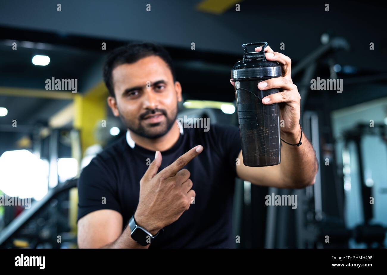 focus on bottle, Confident indian bodybuilder shacking protein bottle and showing by looking at camera - concept of healthy lifestyle, advertisement Stock Photo