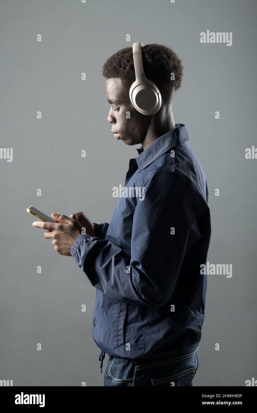 Side view of confident young African American male millennial in trendy outfit and headphones using mobile phone while listening to music against gray Stock Photo