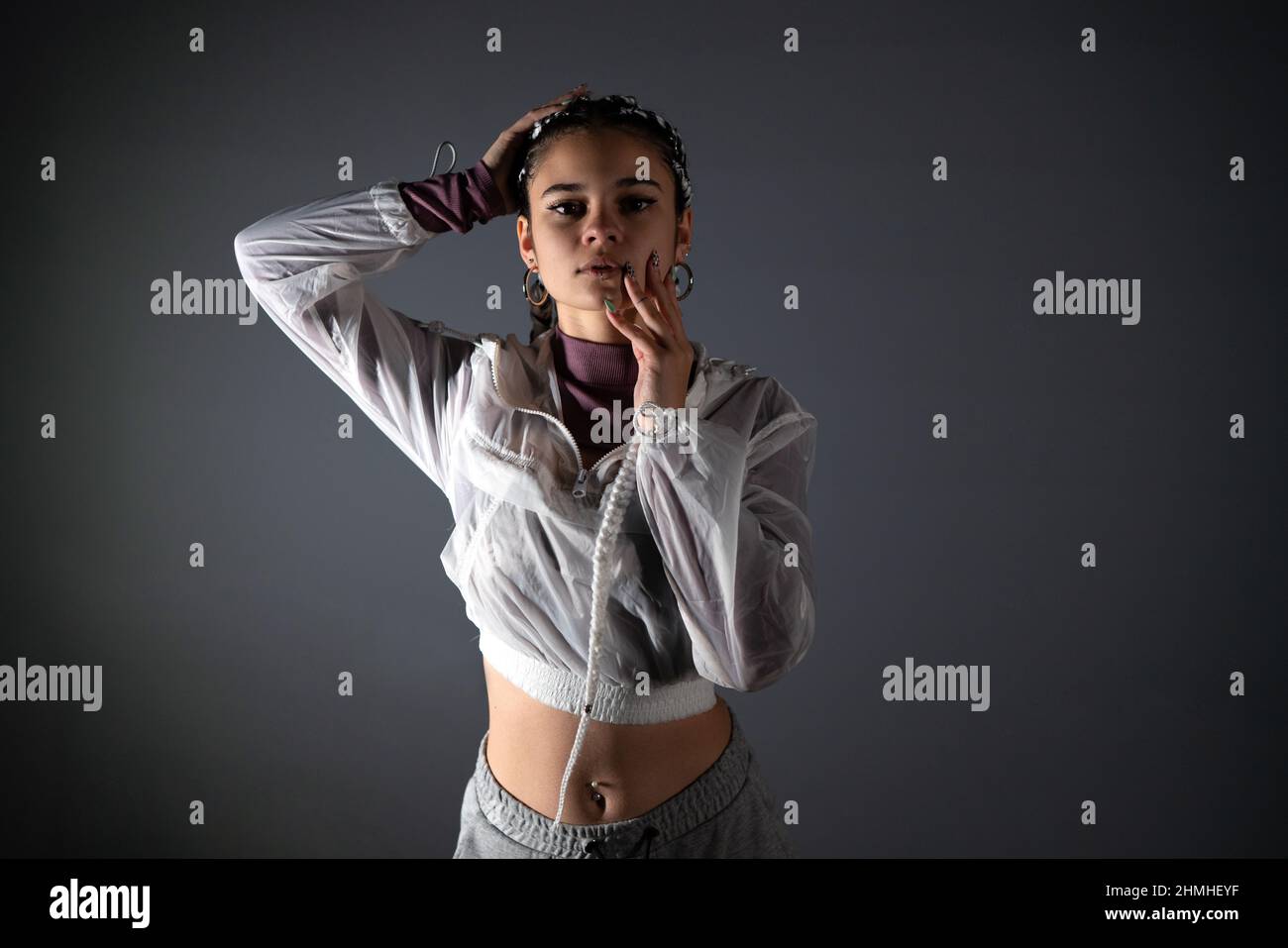 Front view of a young woman with urban style looking at camera while posing with her hands on her face and hair in studio shot. Stock Photo