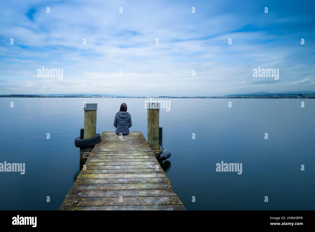 Self reflection. One woman sits on a wooden pier. Cloudy above the lake. Long exposure. Stock Photo