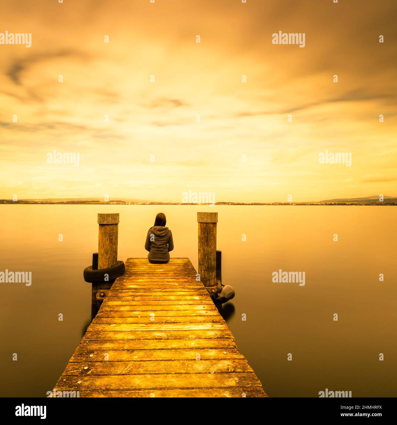 Self reflection in magical world of fantasy. One woman sits on a wooden pier. Cloudy above the lake. Long exposure. Stock Photo