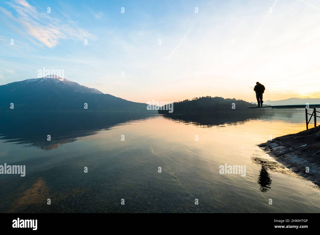 A man catches a fish. Happy relaxed hours alone with nature. Landscape of a mountain lake. Evening light of the setting sun. Male silhouette with a fi Stock Photo