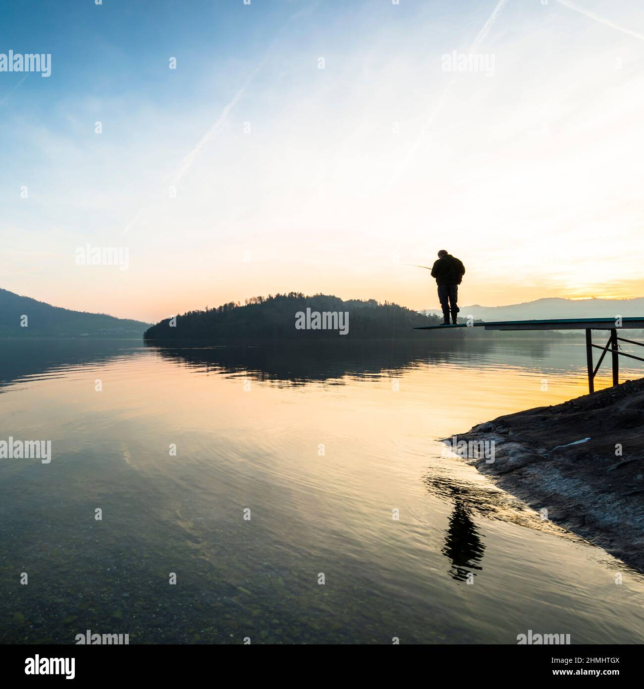 A man catches a fish. Happy relaxed hours alone with nature. Landscape of a mountain lake. Evening light of the setting sun. Male silhouette with a fi Stock Photo