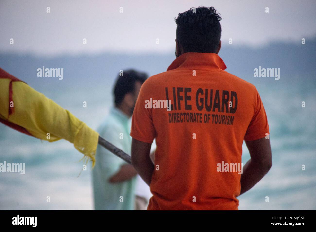 lifeguard in orange shirt carrying flag along the beach with tourists in background at havelock swaraj dweep andaman nicobar islands india Stock Photo
