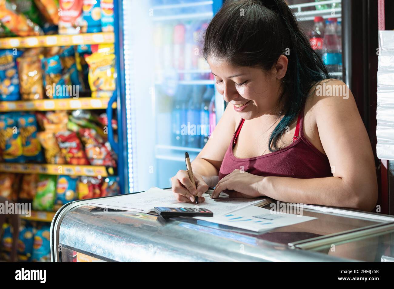 beautiful latin woman smiling very happy, writing in her notebook the excellent performance of her grocery store, girl leaning on glass display case v Stock Photo