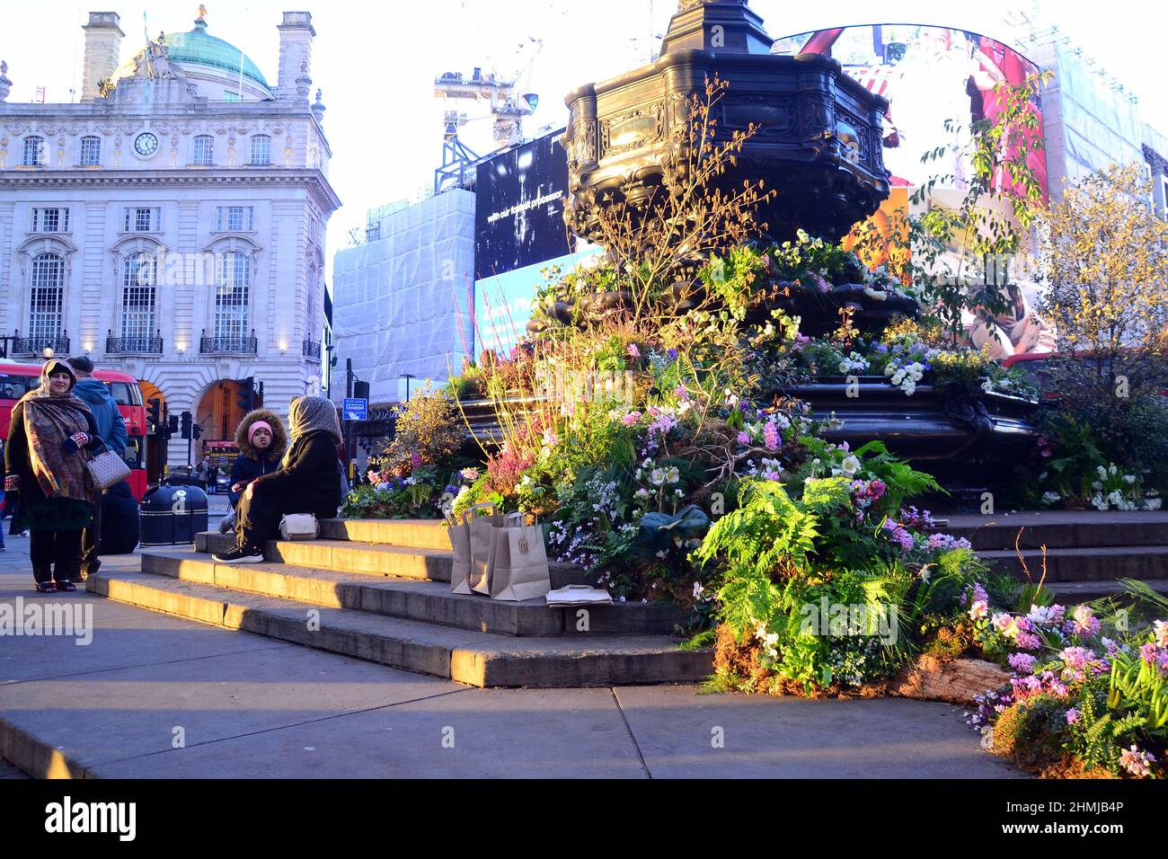 Plants and flowers cover the Shaftesbury Memorial Fountain,  often known as 'Eros', in Piccadilly Circus, London, England, United Kingdom. This advertises  a Green Planet augmented reality experience opening in Piccadilly Circus on 11th February, 2022. Partners and supporters in delivering the project include   BBC Earth, EE, Crown Estate, Factory 42, Kew Royal Botanic Gardens, Talesmith and Dimension. The project offers booking of free tickets online and runs until 9th March, 2022. People sit on the statue and enjoy the late afternoon sunshine. Stock Photo