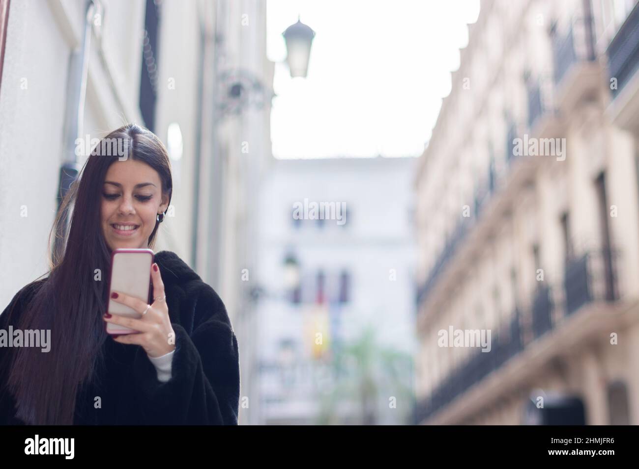 Smiling girl on the street in a black coat holding a mobile phone, typing messages or on a video call. With copy space Stock Photo