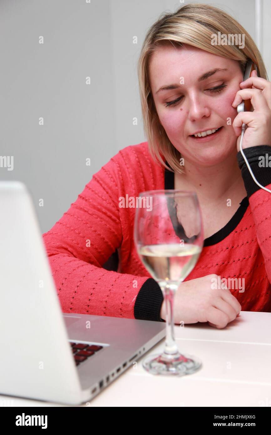 A young female working from home on her laptop and mobile phone, while drinking wine. Stock Photo