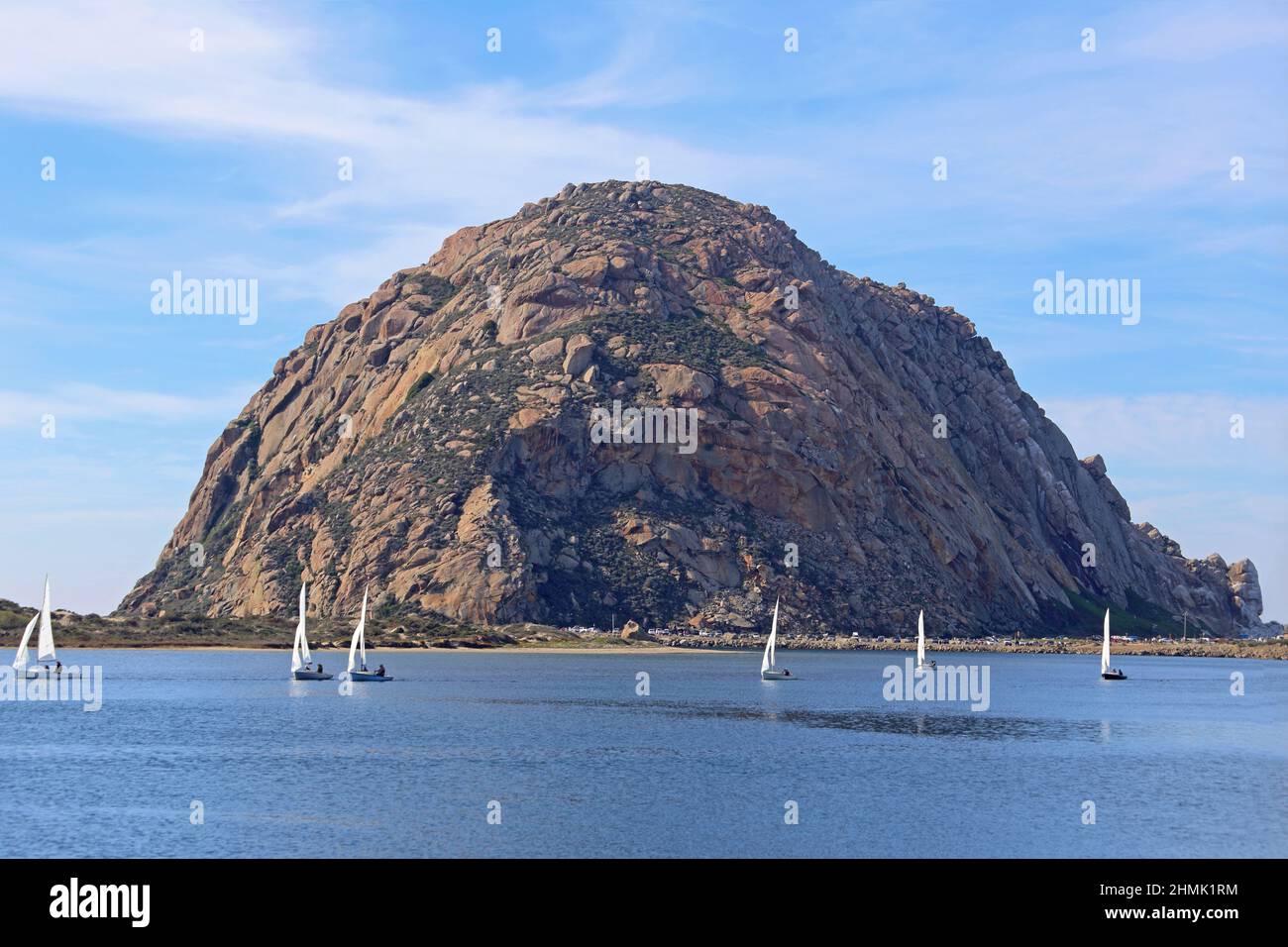 Morro Rock, which is an ancient volcanic plug located in Morro Bay, California, is shown during the day, with dinghy sailboats in the harbor. Stock Photo