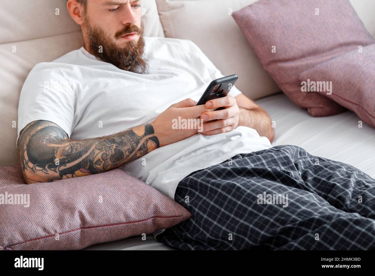 Man in pajamas using smartphone while lying in bed. Young caucasian bearded brutal with tattoos man writes message on mobile phone or surfing in Stock Photo