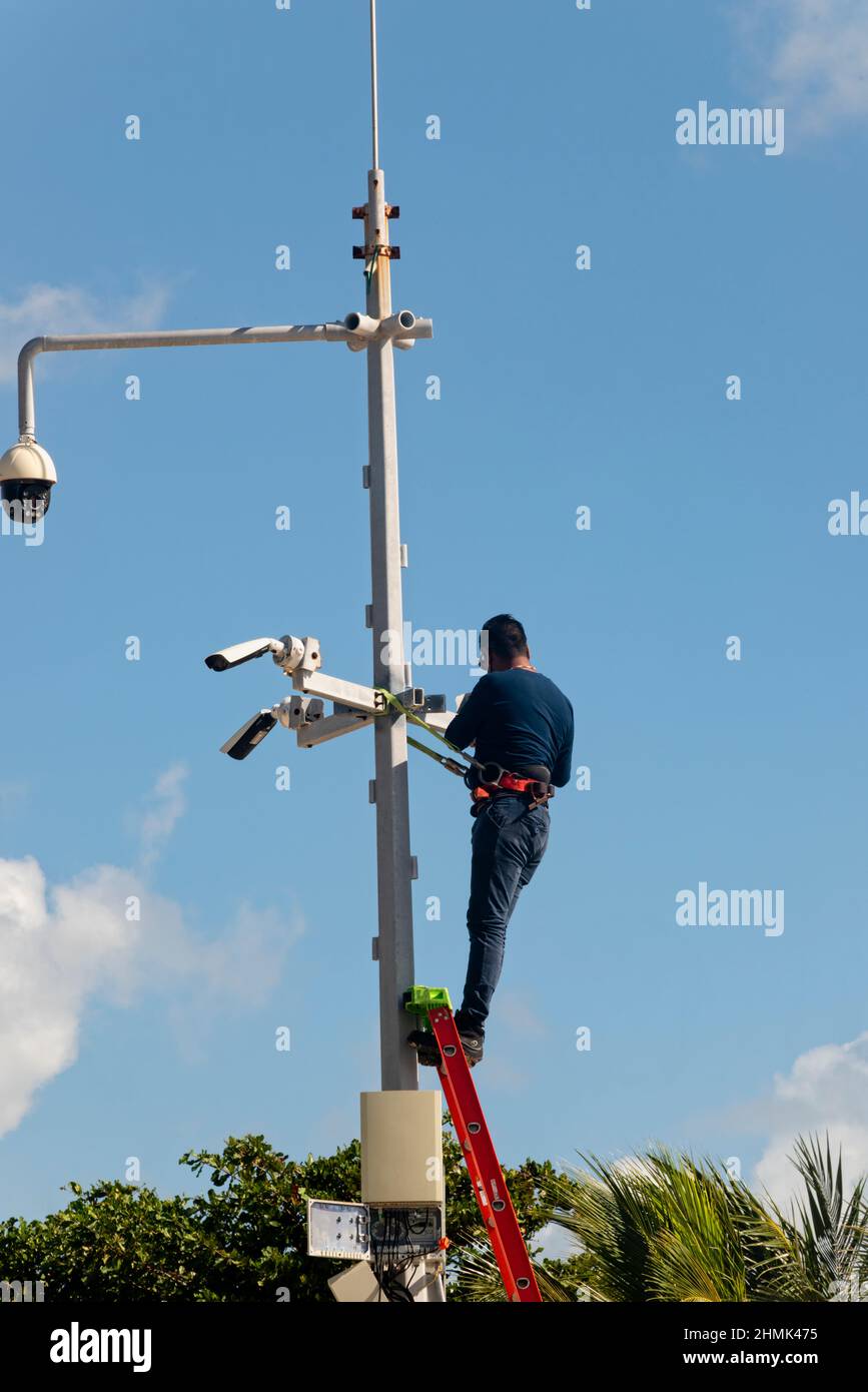 a male worker on a metal ladder installs security  cameras near a public beach in Mexico Stock Photo