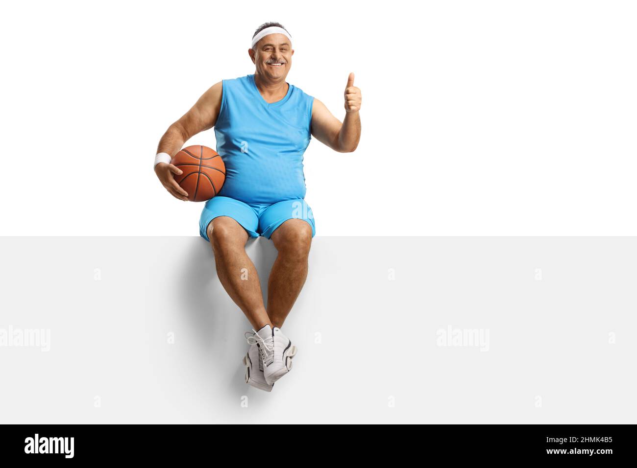 Mature man in a sport jersey with a basketball sitting on a blank panel and gesturing thumbs up isolated on white background Stock Photo
