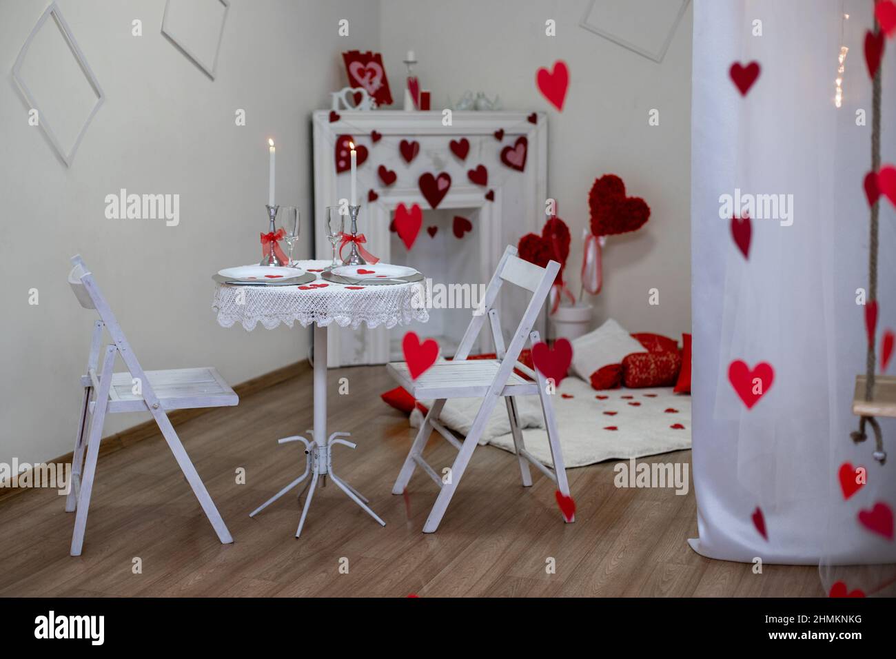 Table with white tablecloth. Romantic candlelight dinner. White fireplace decorated with red hearts for Valentine's Day Stock Photo
