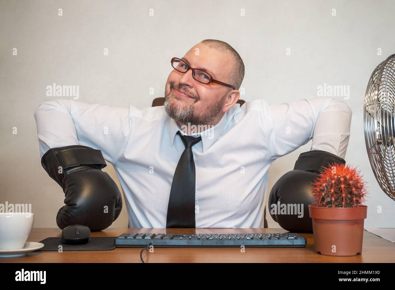 The man comically portrays the work of a businessman. He's sitting at a table wearing boxing gloves. Stock Photo
