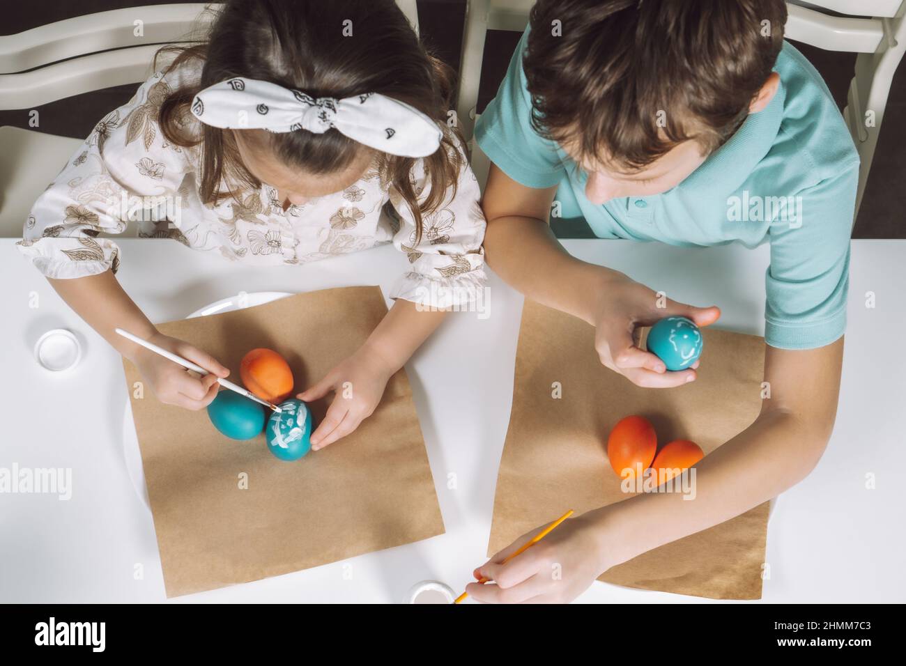 Portrait of two children, little girl and boy siblings, painting multi coloring Easter hens eggs in kitchen, with paintbrushes, sitting chair, paper t Stock Photo