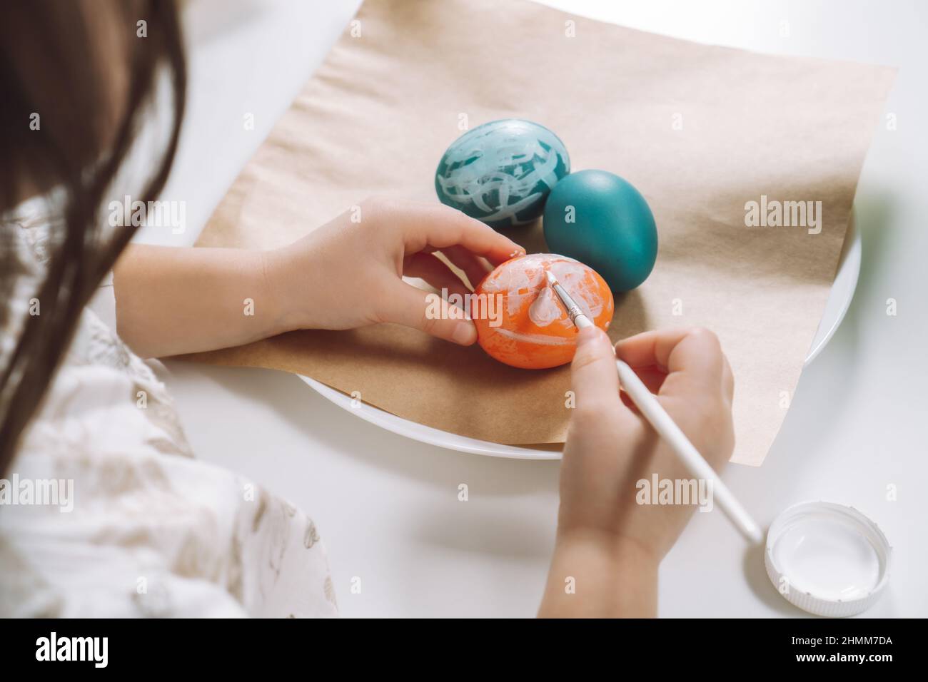 Cropped portrait of girl hand, painting multi coloring Easter hens eggs on brown paper plate table with paintbrush. Making art design white patterns o Stock Photo