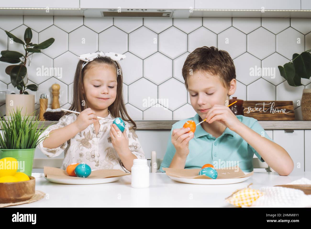 Portrait of concentrated smiling two kids, little girl and boy, coloring patterns on Easter hens eggs on paper table with paintbrushes near plant. Mak Stock Photo