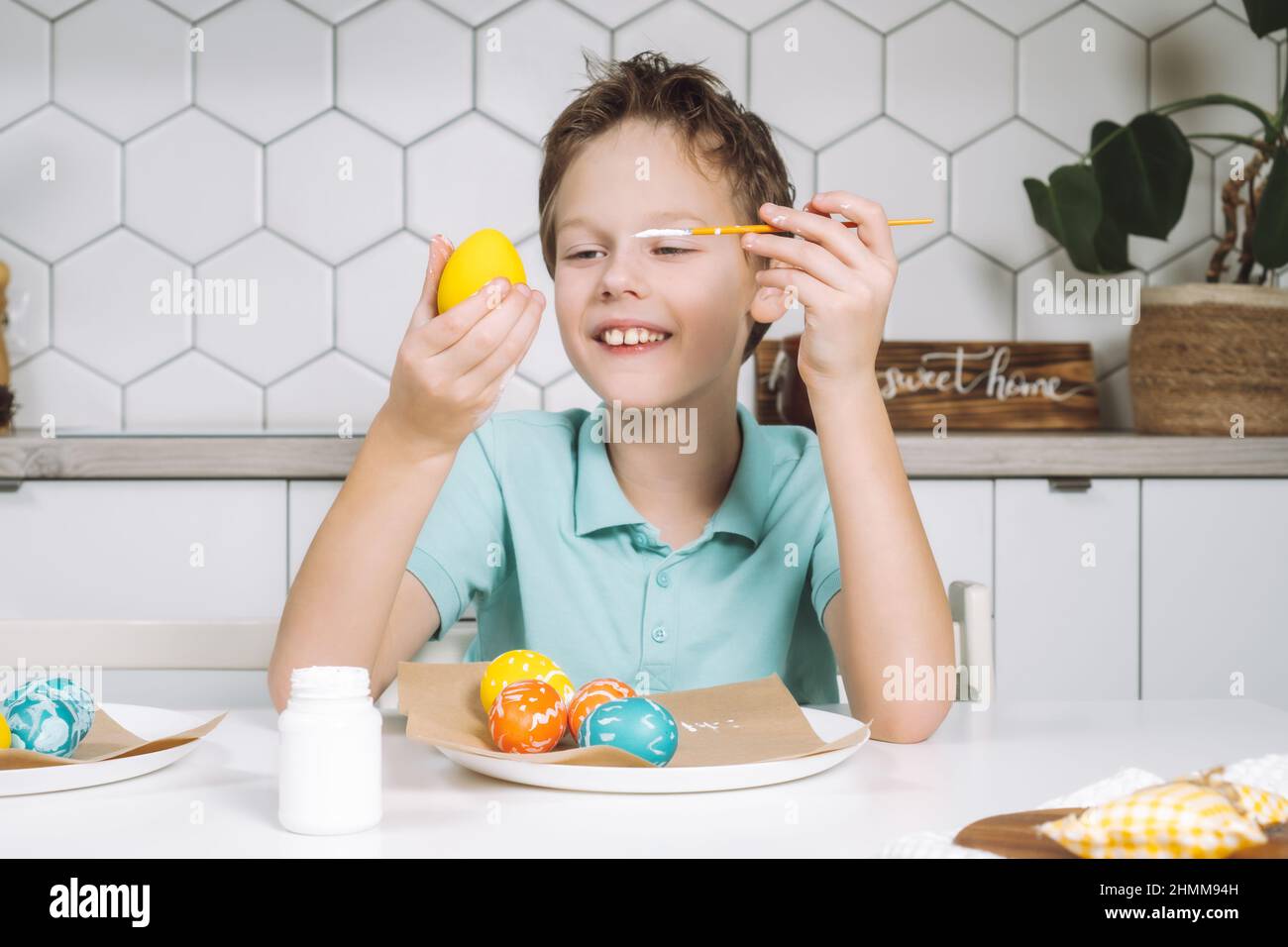 Portrait smiling joyful little boy, painting multi coloring Easter hens eggs paper table with paintbrush. Making art design white patterns. Sitting ch Stock Photo