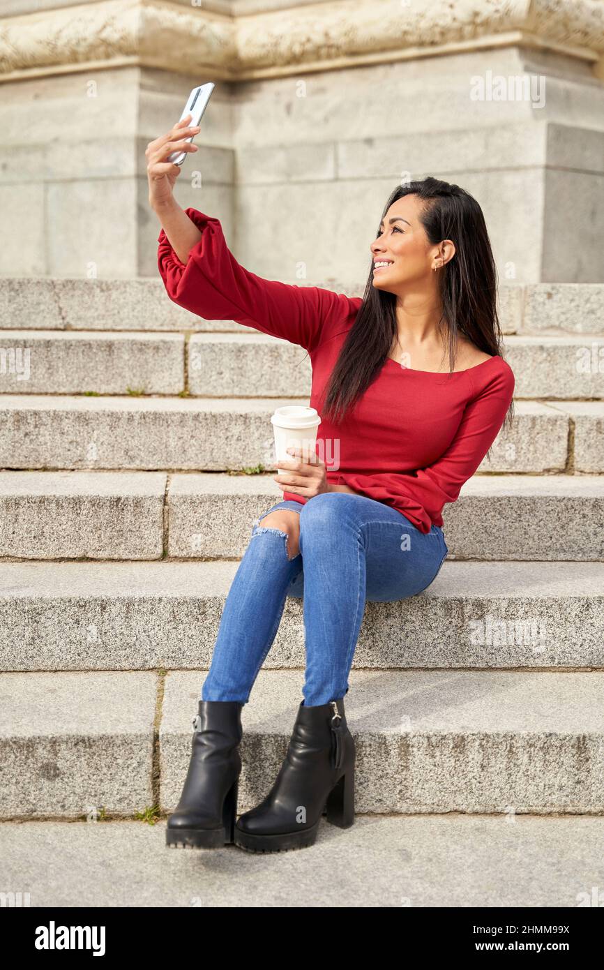 Young Latina girl taking a selfie with cell phone, relaxing on stairs on a spring day. Latina in red sweater and jeans relaxing in the city. Lifestyle concept.  Stock Photo
