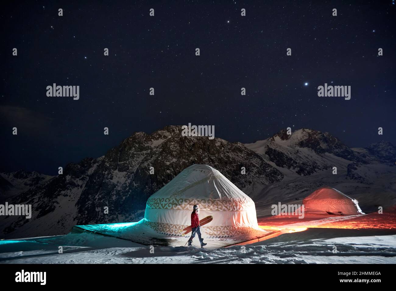 A man with a snowboard walking near a yurt at night in winter in the mountains Stock Photo