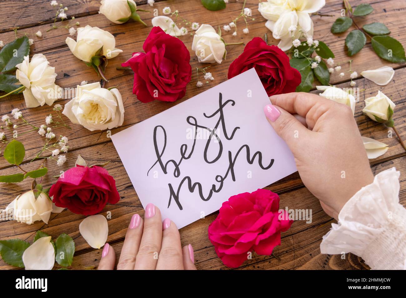 https://c8.alamy.com/comp/2HMMJCW/hands-with-handwritten-card-best-mom-surrounded-by-red-and-cream-roses-close-up-on-a-wooden-table-femminine-romantic-declaration-of-love-near-flowers-2HMMJCW.jpg