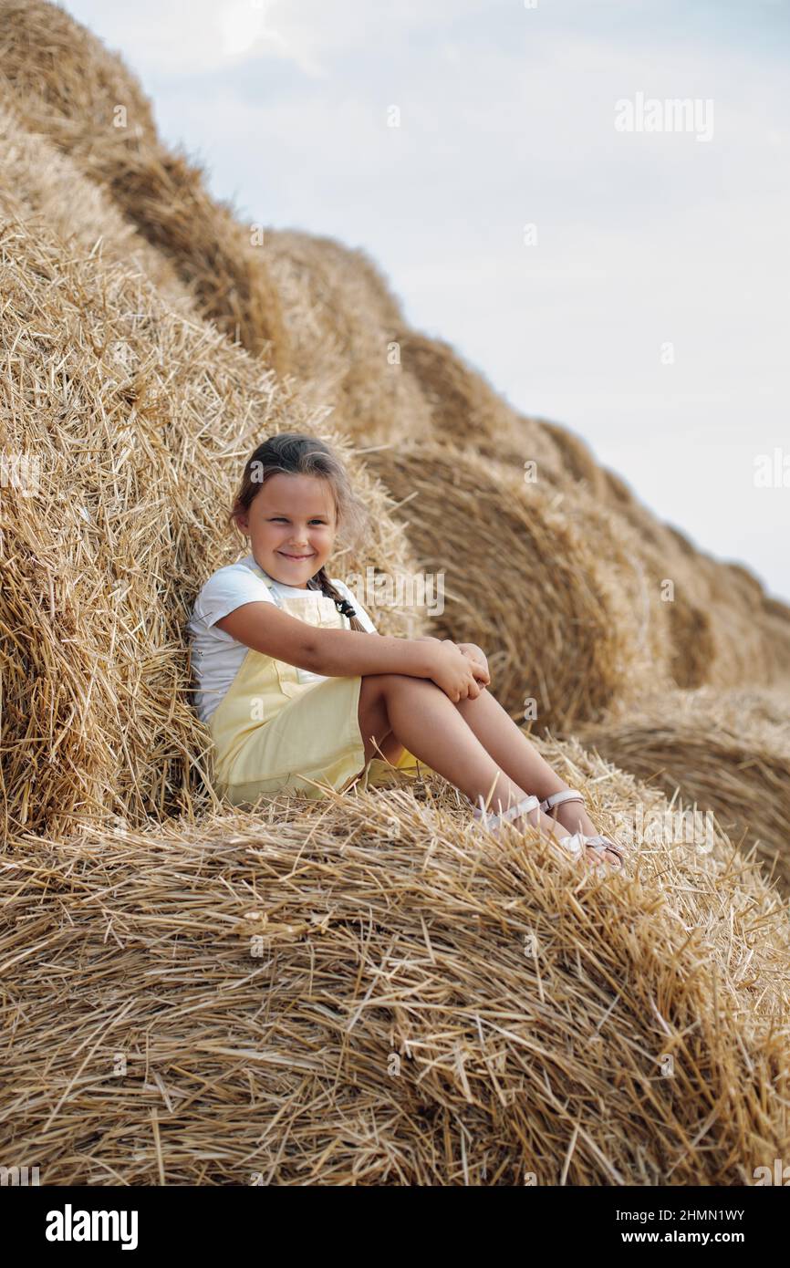 Portrait of sly small girl resting on haystack looking at camera with squinted eyes wearing sundress with dozens of haystacks in background. Away from Stock Photo