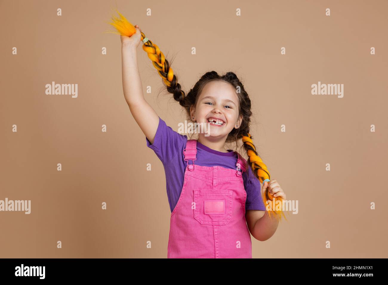 Joyful excited young girl playing with yellow kanekalon braids, having big smile with missing tooth looking at camera wearing pink jumpsuit and purple Stock Photo